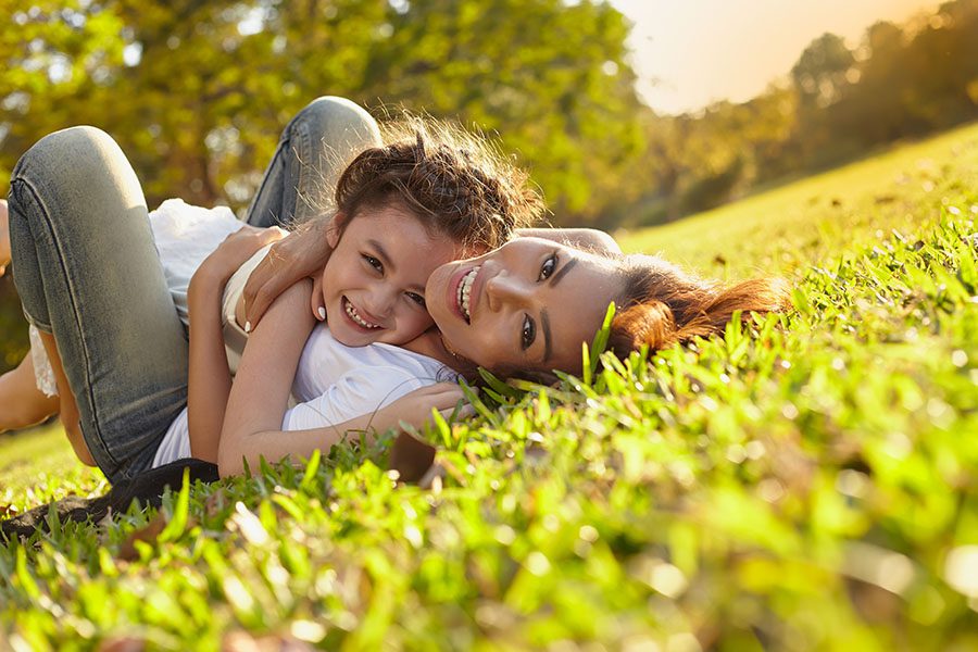 About Our Agency - Closeup Portrait of a Cheerful Mother and Daughter Laying in the Green Grass and Having Fun Playing Together On a Warm Summer Day