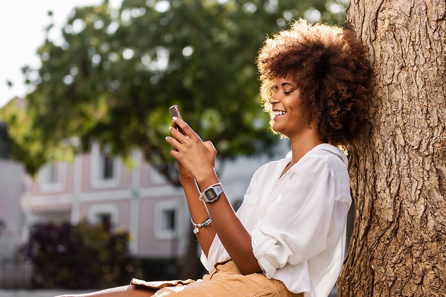 Contact - Closeup Portrait of a Cheerful Young Woman Sitting Next to a Tree in a Quiet Residential Neighborhood in the City While Using a Phone